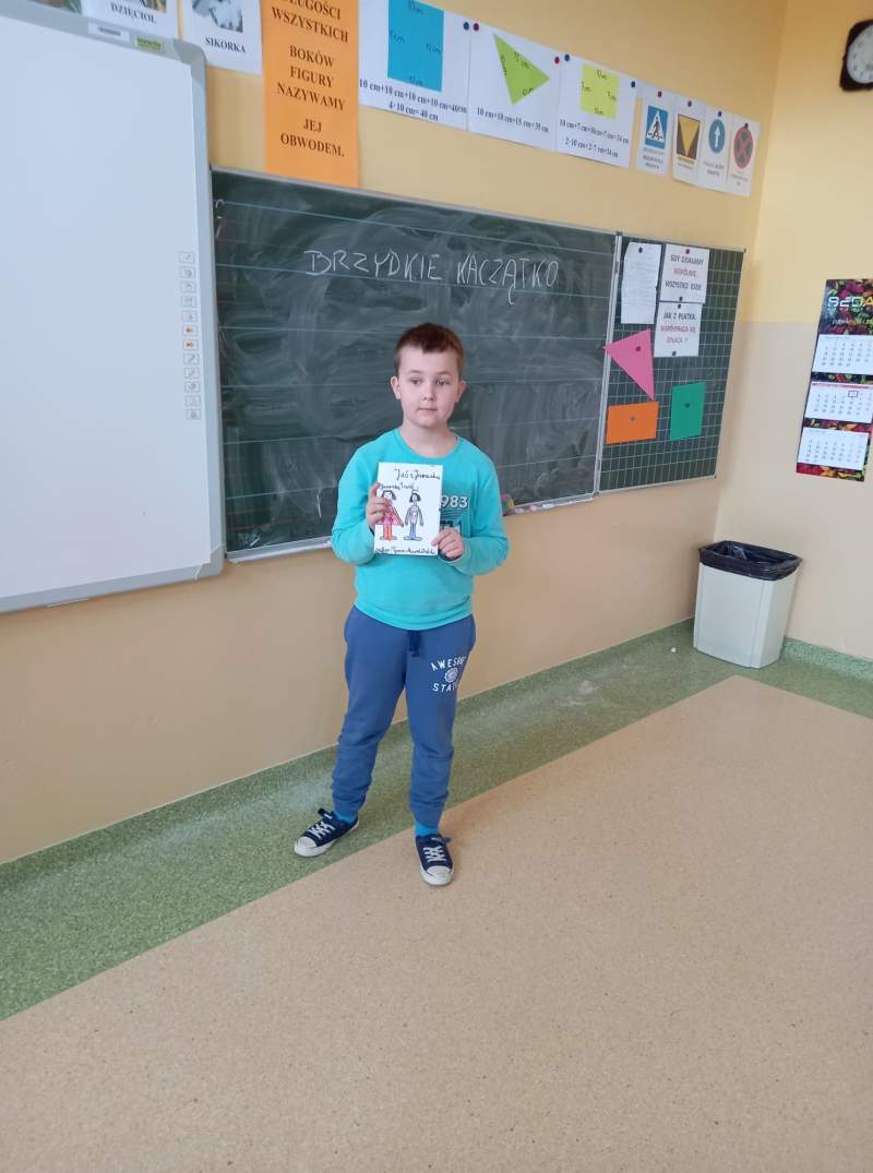 Fotografia przedstawia ucznia stojącego w klasie przy tablicy. Uczeń trzyma w ręce wykonana przez siebie pracę plastyczną