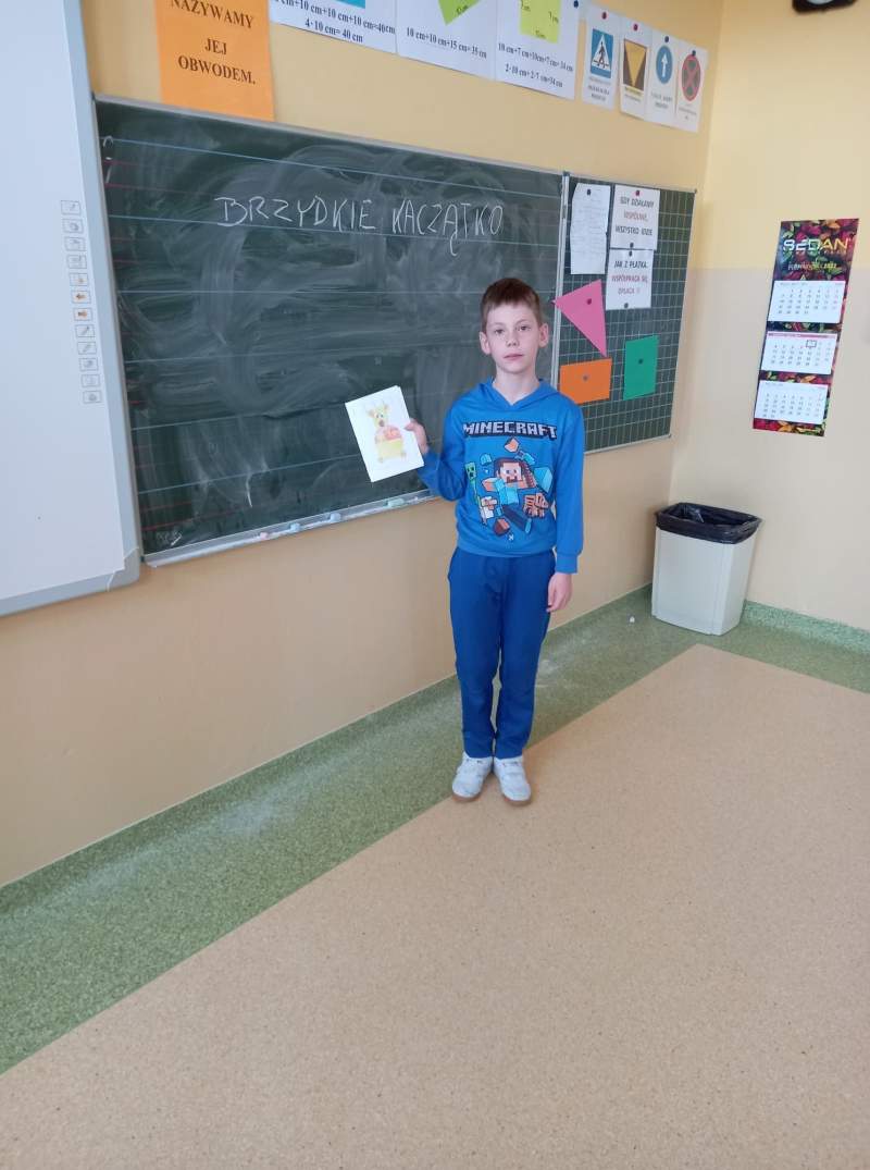 Fotografia przedstawia ucznia stojącego w klasie przy tablicy. Uczeń trzyma w ręce wykonana przez siebie pracę plastyczną