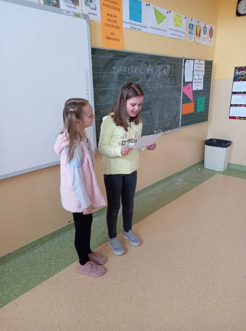 Fotografia przedstawia dwie uczennice stojące w klasie przy tablicy. Uczennice czytają napisaną przez siebie bajkę