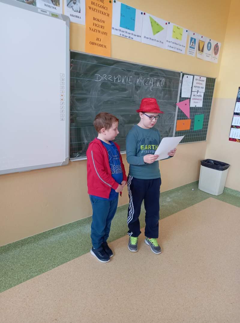 Fotografia przedstawia dwóch uczniów stojących w klasie przy tablicy. Uczniowie czytają napisaną przez siebie bajkę