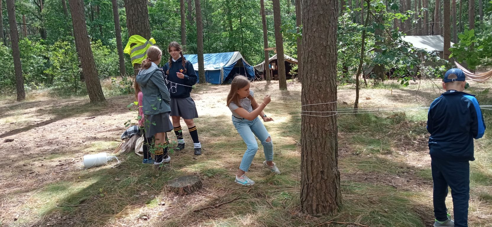 Fotografia przedstawia dzieci bawiących się z harcerzami w lesie