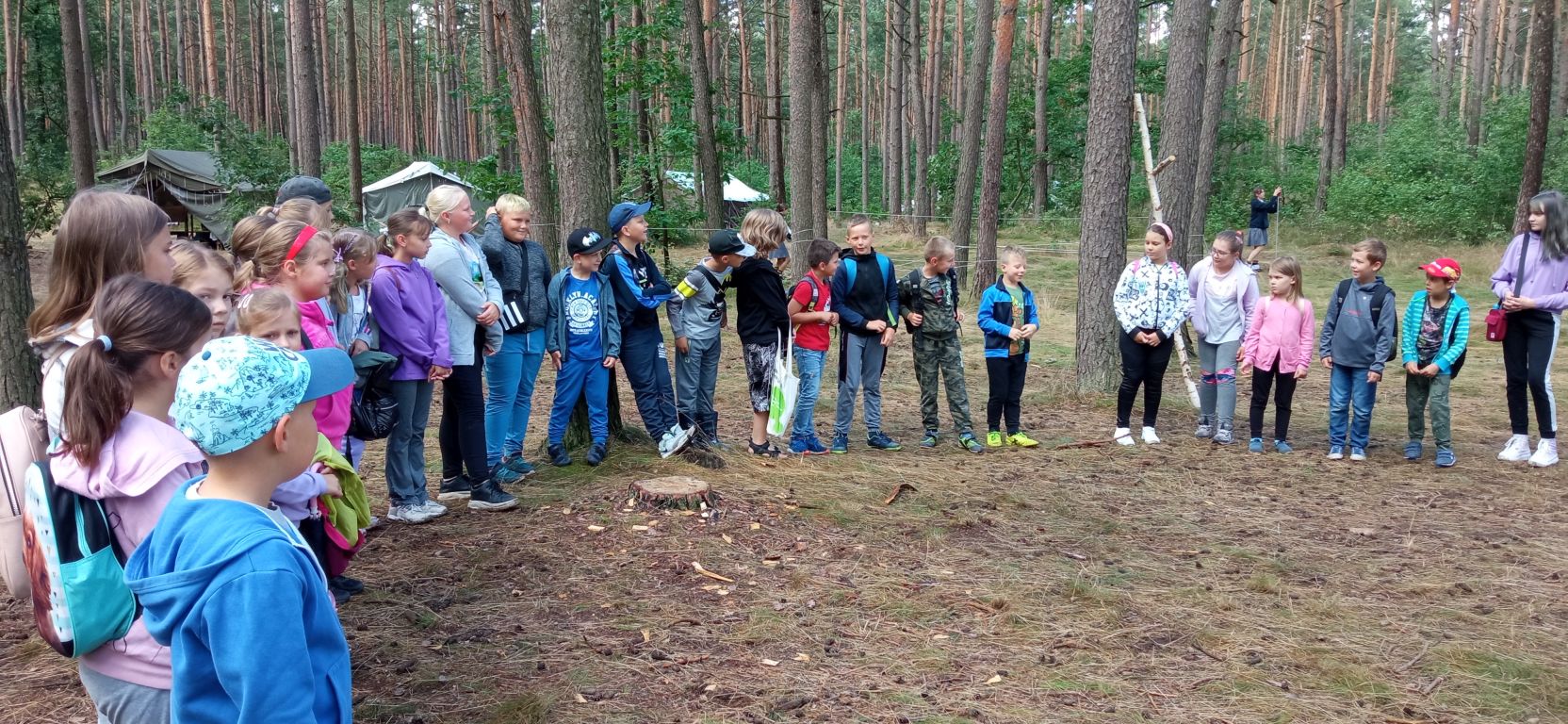 Fotografia przedstawia grupę uczestników zajęć wakacyjnych stojących w kręgu w lesie na zajęciach z harcerzami