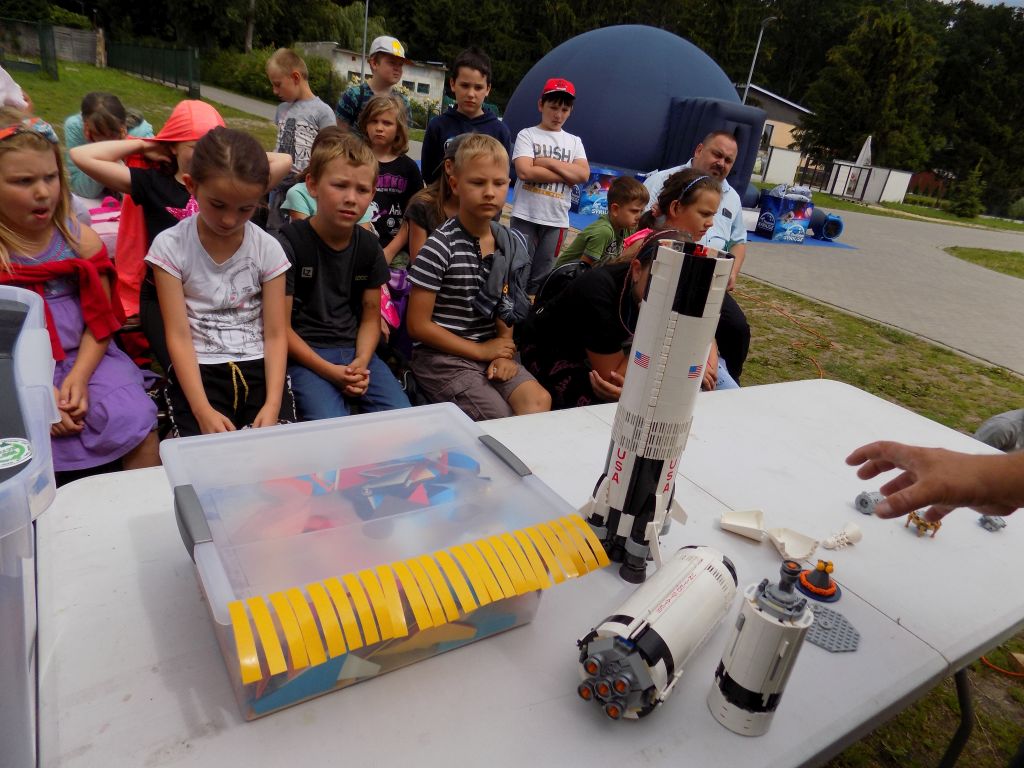 Fotografia przedstawia uczestników zajęć obserwujących budowę rakiety rozłożonej na części. Obok na stole stoi organizer, a w tle widać namiot