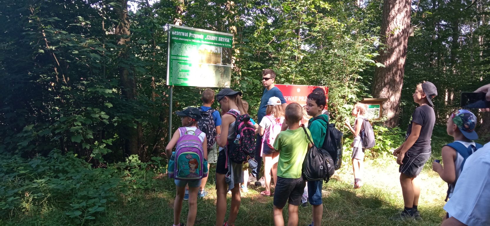 Fotografia przedstawia uczestników zajęć z przewodnikiem stojących na terenie rezerwatu przyrody Czarny Bryńsk. Dzieci słuchają wypowiedzi przewodnika na temat rezerwatu