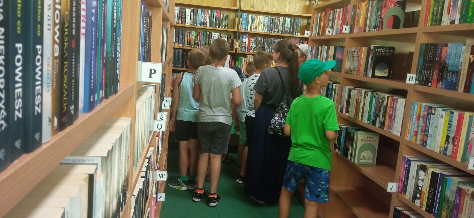 Fotografia przedstawia uczestników zajęć w bibliotece. Dzieci krzątają się między regałami