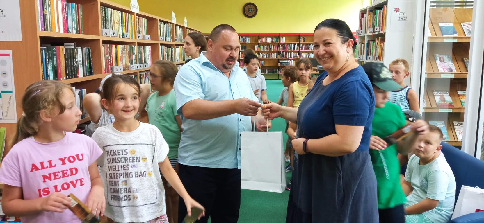 Fotografia przedstawia Dyrektora Biblioteki wręczającego upominki jednej z pracownic biblioteki, a w tle radosne dzieci