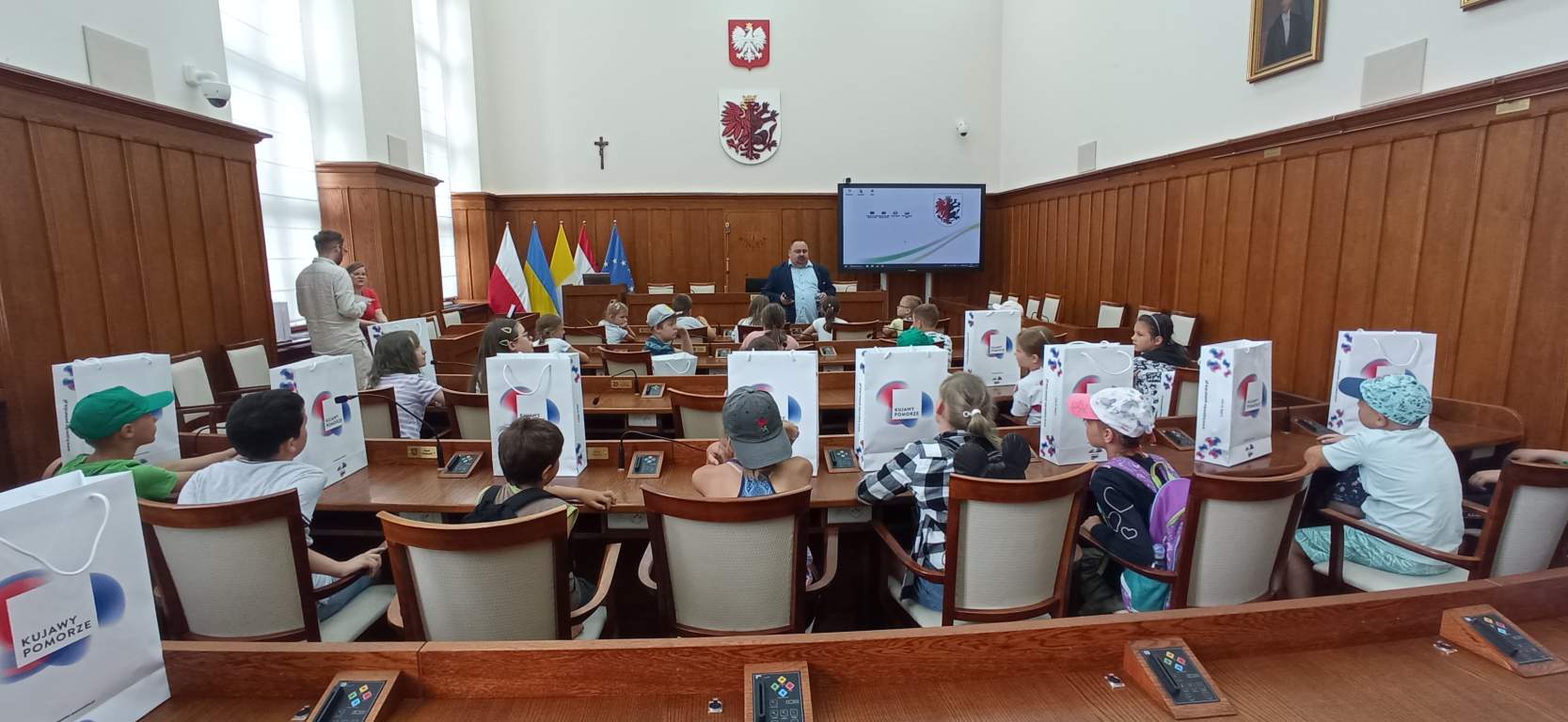 Fotografia przedstawia uczestników zajęć wraz z Dyrektorem Biblioteki w Sejmiku Wojewódzkim. Słuchają wypowiedzi naszego dyrektora a w tle widać flagi i godło