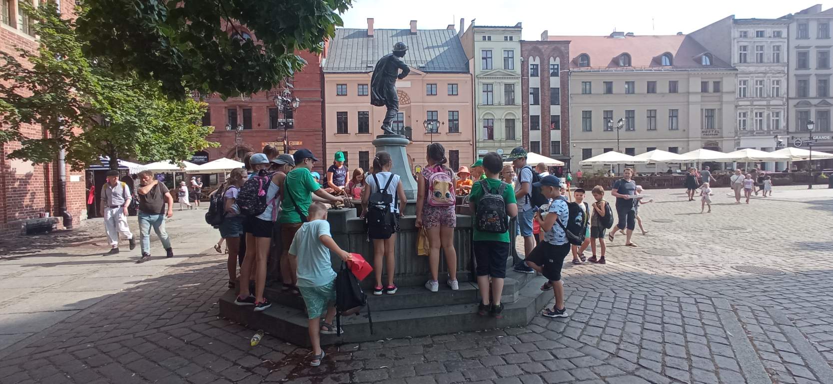 Fotografia przedstawia uczestników zajęć przy fontannie na Starówce w Toruniu. W tle widać starówkę