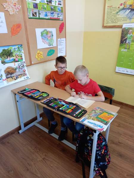 Na zdjęciu są dwaj chłopcy siedzący przy ławce. Jeden z nich czyta książkę, a drugi słucha. Na ścianie obok nich wisi duża tablica ozdobiona jesiennymi elementami
