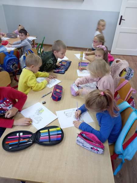 Na zdjęciu widać dzieci, które kolorują rysunki związane z dniem głośnego czytania. Siedzą przy stolikach, a na nich leżą piórniki, w których są kredki.