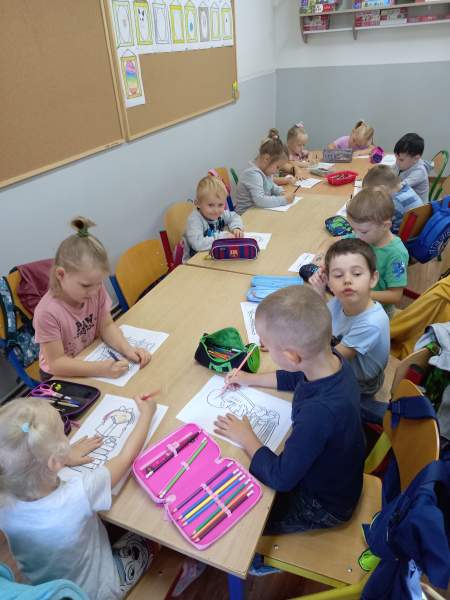 Zdjęcie przedstawia dzieci siedzące przy trzech połączonych ze sobą stolików. Kolorują swoje prace
