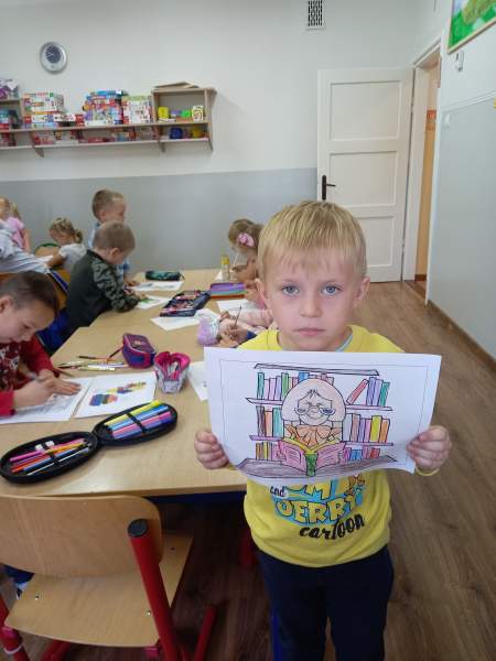 Zdjęcie przedstawia chłopca dumnie prezentującego swój pokolorowany rysunek. Rysunek przedstawia kobietę czytającą książkę w bibliotece. W tle inne dzieci, które również kolorują swoje rysunki