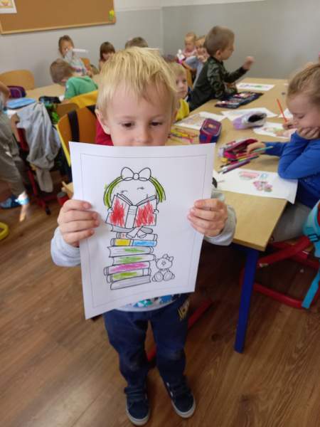 Zdjęcie przedstawia chłopca, który prezentuje swoją pracę. Na rysunku jest dziewczynka siedząca na kolumnie książek