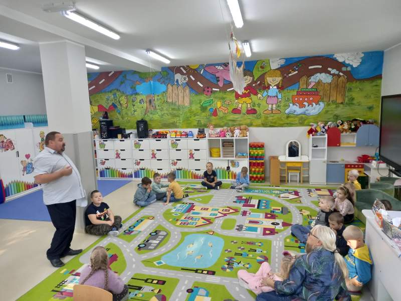 Na zdjęciu widać dzieci, ich opiekuna oraz dyrektora biblioteki w Skępem. Siedzą w przedszkolu we Wiosce. Słuchają opowieści dyrektora