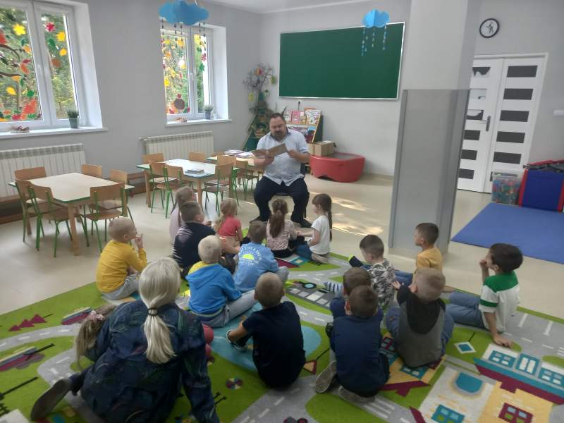Na zdjęciu widać siedzące dzieci słuchające bajki czytanej przez dyrektora. Na szybach okien przyklejone są jesienne ozdoby