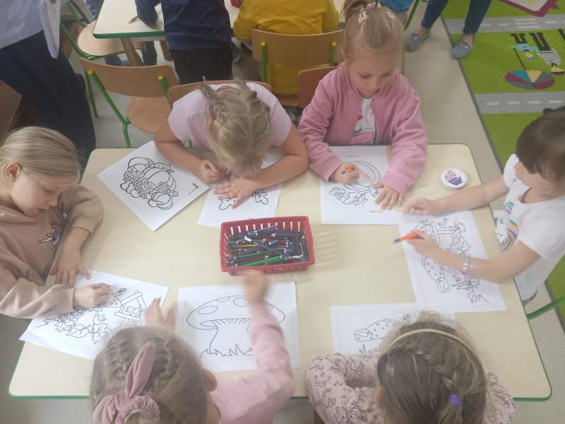Na zdjęciu są dzieci kolorujące tematyczne rysunki nawiązujące do jesieni. Siedzą wspólnie przy stole, a na środku leży pudełko z kredkami