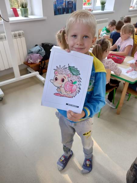 Na zdjęciu widać chłopca prezentującego swój rysunek, który przedstawia jeża. W tle są dzieci, które dalej kolorują swoje prace