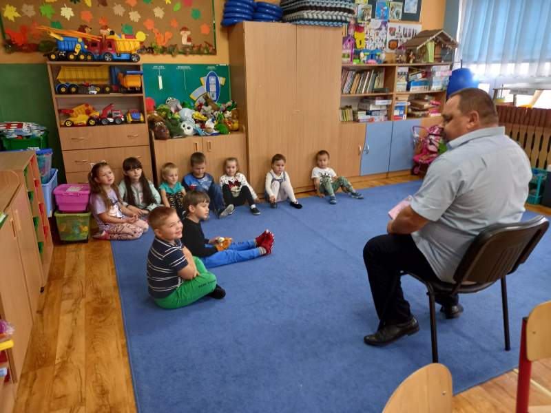 Na zdjęciu widać dzieci siedzące na wykładzinie, które dyskutują z Dyrektorem biblioteki siedzącym na krześle