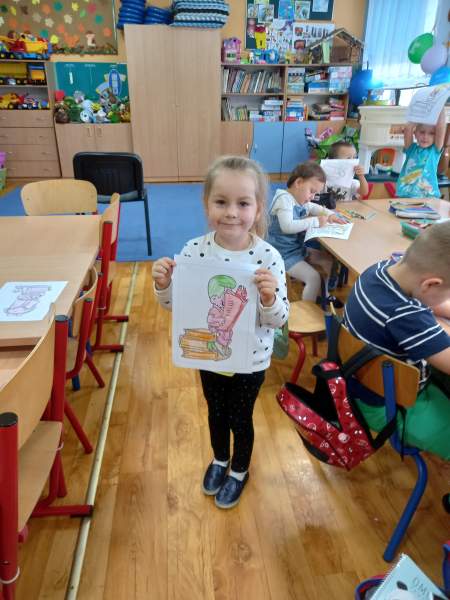 Na zdjęciu widać dziewczynkę uczestniczącą w zajęciach, która dumnie pokazuje do zdjęcia swój pokolorowany rysunek. W tle dzieci, które dalej kolorują swoje prace