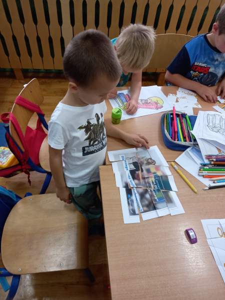 Na zdjęciu widać trzech chłopców aktywnie biorących udział w zajęciach. Układają tematyczne puzzle i kolorują. Na stole rozłożone są kredki