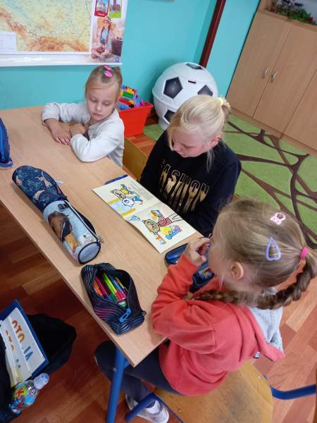 Na zdjęciu widać trzy dziewczynki przy stoliku. Dziewczynki czytają wspólnie książkę o Kici Koci