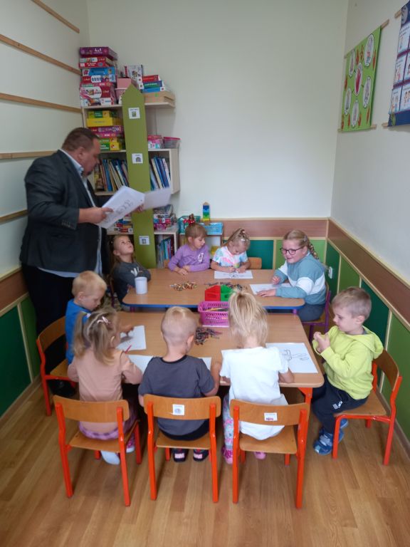 Zdjęcie przedstawia dyrektora wręczającego dzieciom siedzącym na krzesłach przy stoliku w przedszkolu kolorowanki