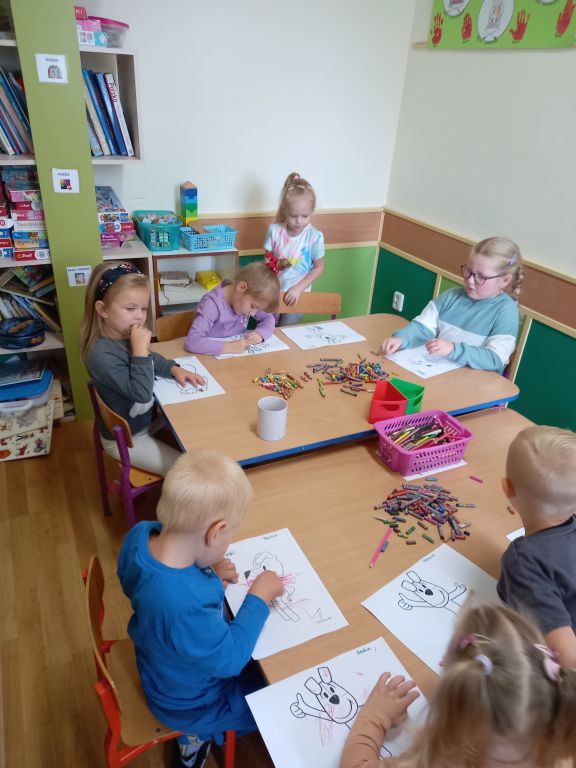 Zdjęcie przedstawia dzieci siedzące na krzesłach przy stoliku w trakcie kolorowania obrazków