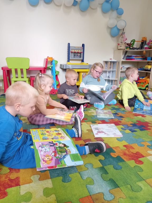 Zdjęcie przedstawia dzieci siedzące w przedszkolu na dywanie i oglądające kolorowe książeczki