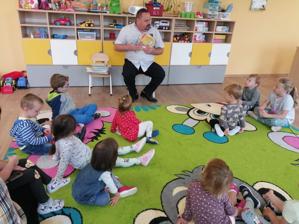 Dyrektor biblioteki siedzi na krześle w przedszkolu, na dywanie przed nim siedzą dzieci. Dyrektor pokazuje dzieciom kolorową książeczkę