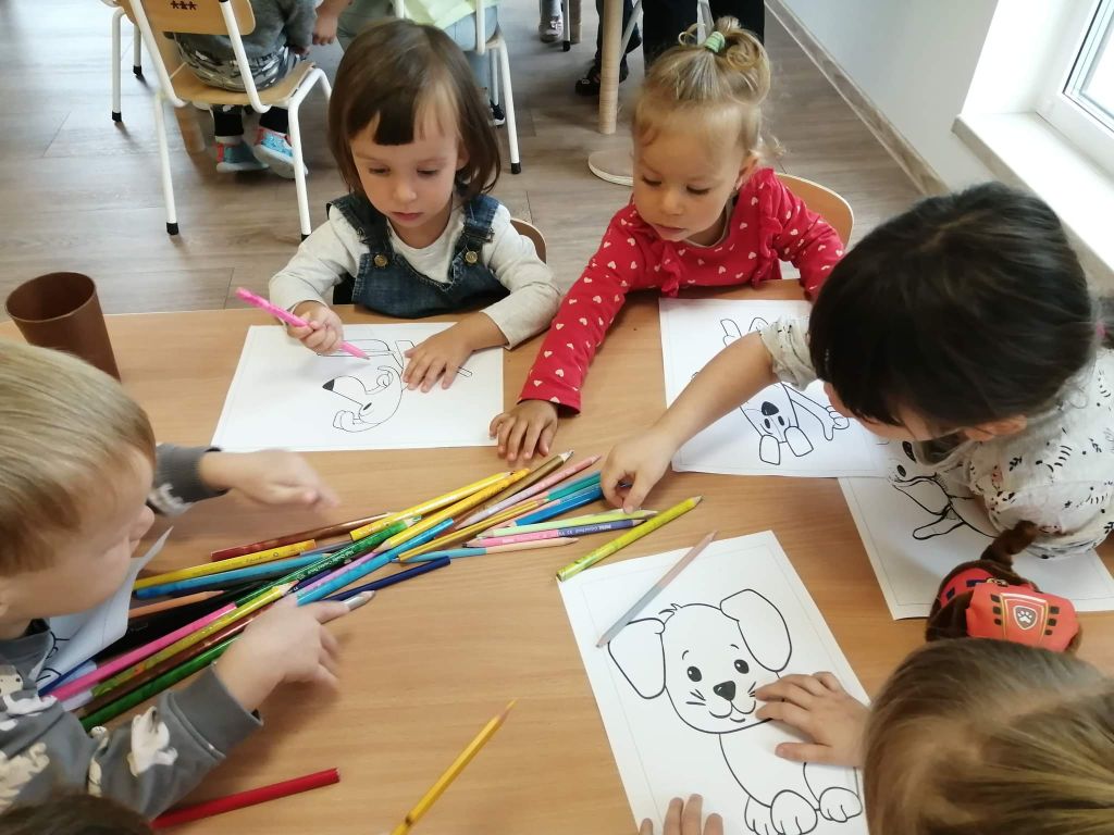 Fotografia przedstawia dzieci w przedszkolu siedzące przy stoliku i kolorujące kredkami obrazki