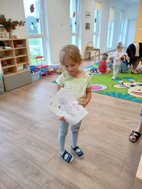 Fotografia przedstawia dziewczynkę stojącą w przedszkolu i trzymajacą w rękach pokolorowany obrazek Reksia