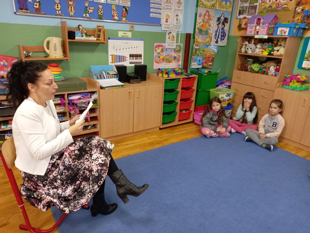 Fotografia przedstawia nauczycielkę czytającą dzieciom wiersz w klasie 