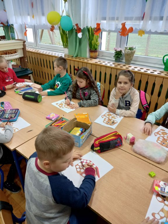 Fotografia przedstawia dzieci siedzące w szkolnych ławkach w trakcie lekcji kolorujące szablony na kartkach papieru