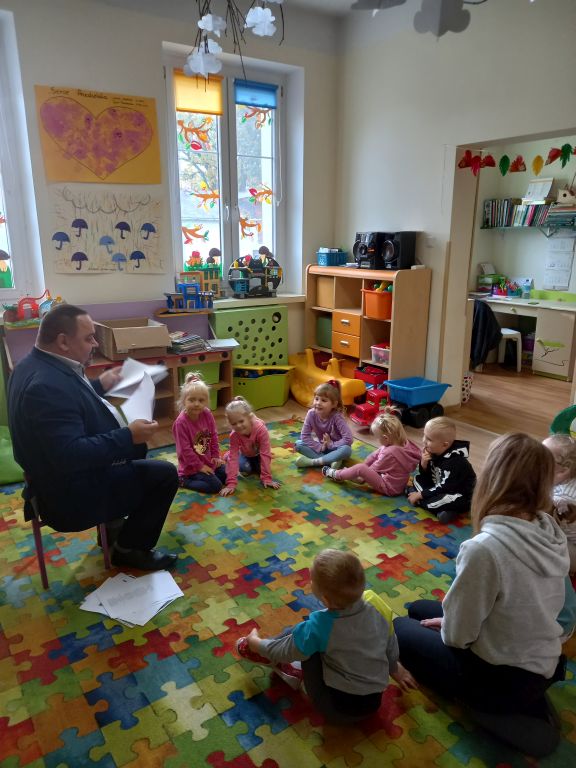 Fotografia przedstawia Dyrektora Biblioteki siedzącego na krześle w przedszkolu. Dyrektor trzyma w rękach obrazki i pokazuje je dzieciom siedzącym przed nim na dywanie