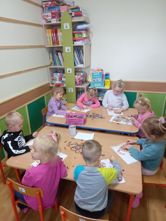 Fotografia przedstawia przedszkolaków siedzących w przedszkolu przy stolikach w trakcie zajęć