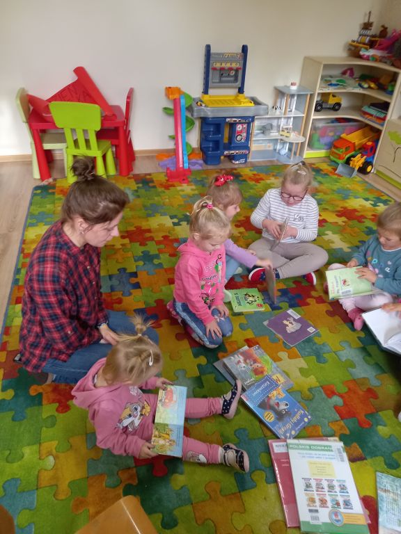 Fotografia przedstawia dzieci i wychowawczynię siedzących na dywanie w przedszkolu. Dzieci oglądają kolorowe książeczki