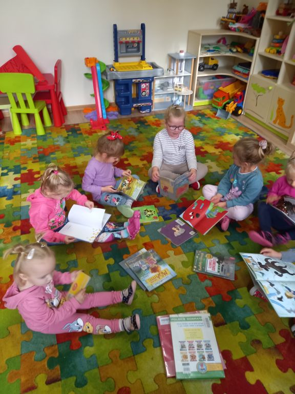 Fotografia przedstawia dzieci siedzące na dywanie w przedszkolu. Dzieci oglądają kolorowe książeczki