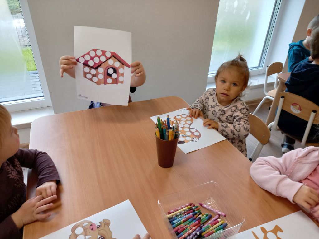 Fotografia przedstawia dzieci siedzące w przedszkolu przy stolikach. Dzieci kolorują obrazki