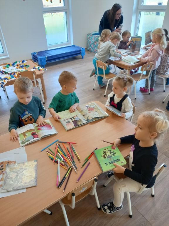 Fotografia przedstawia dzieci siedzącego przy stoliku w przedszkolu i oglądające kolorowe książeczki