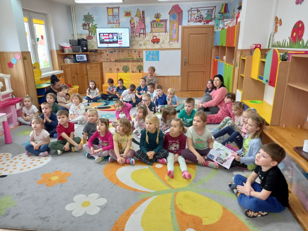Zdjęcie przedstawia dzieci wraz z opiekunami siedzące w przedszkolu na dywanie