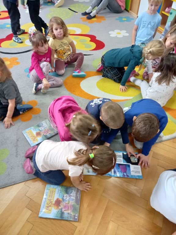 Zdjęcie przedstawia dzieci siedzące na dywanie w przedszkolu. Dzieci oglądają kolorowe książeczki
