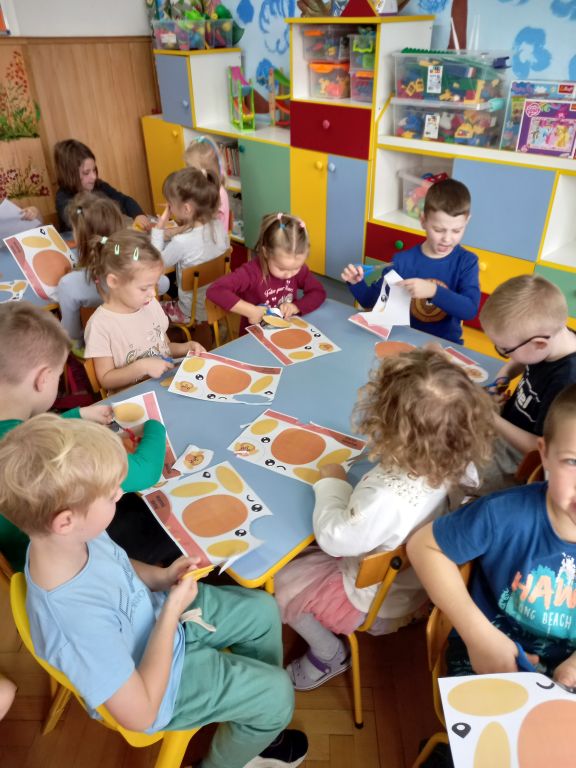 Zdjęcie przedstawia dzieci siedzące w przedszkolu przy stolikach. Dzieci wycinają z szablonów papieru głowę pieska