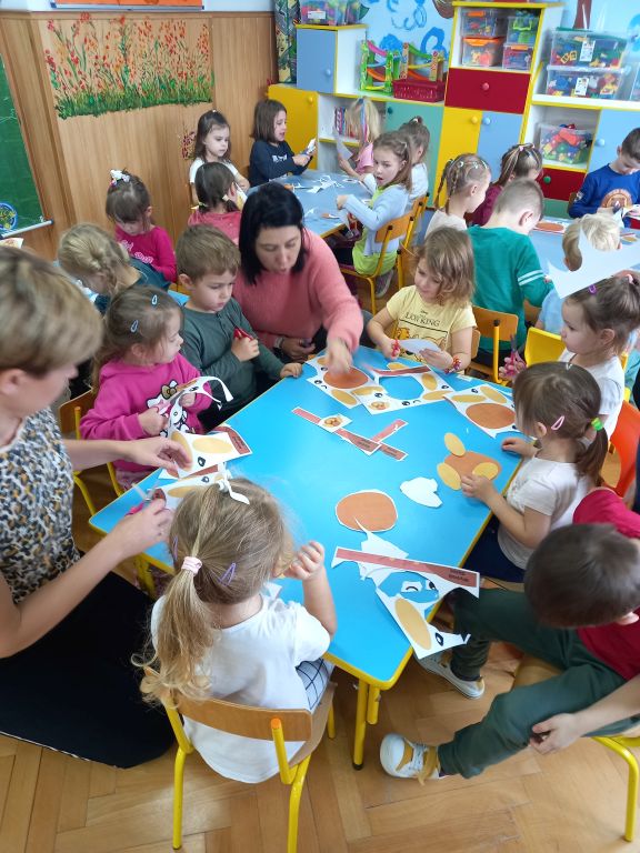 Zdjęcie przedstawia dzieci wraz z opiekunami siedzące w przedszkolu przy stolikach. Dzieci i opiekunowie wycinają z szablonów papieru głowę pieska