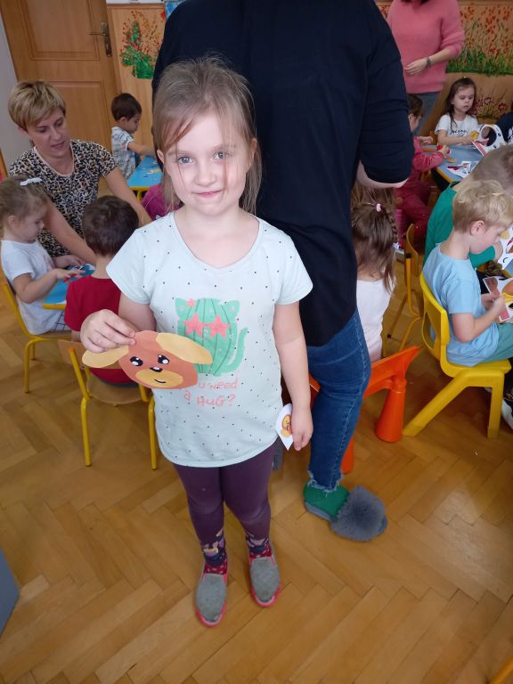 Zdjęcie przedstawia dziewczynkę w przedszkolu. Dziewczynka w rękach trzyma ukończoną pracę plastyczną