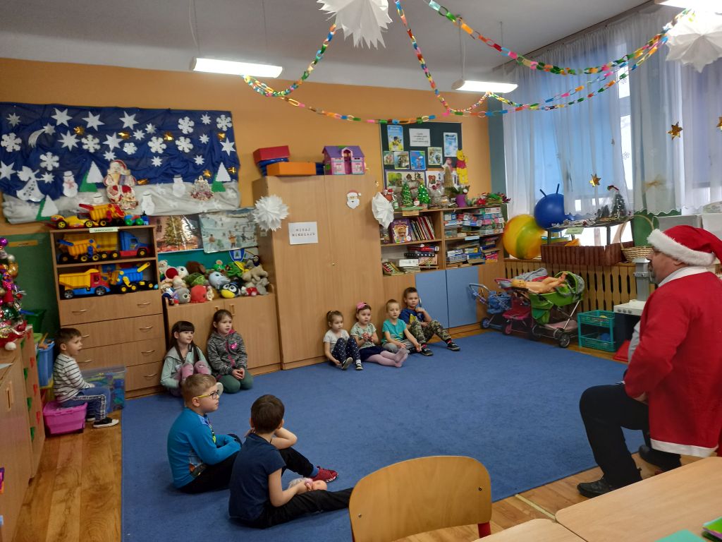 Zdjęcie przedstawia dyrektora biblioteki siedzącego na krześle w przedszkolu w stroju Mikołaja. Wokoło dyrektora siedzą na dywanie dzieci zgłaszające się do odpowiedzi z rękami uniesionymi do góry