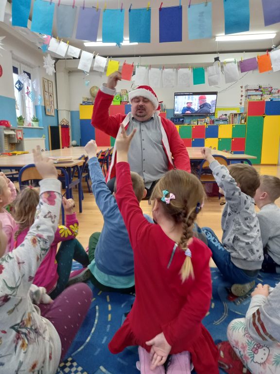 Zdjęcie przedstawia dyrektora biblioteki w przedszkolu w stroju Mikołaja. Dyrektor wręcza dzieciom czekoladowe Mikołaje