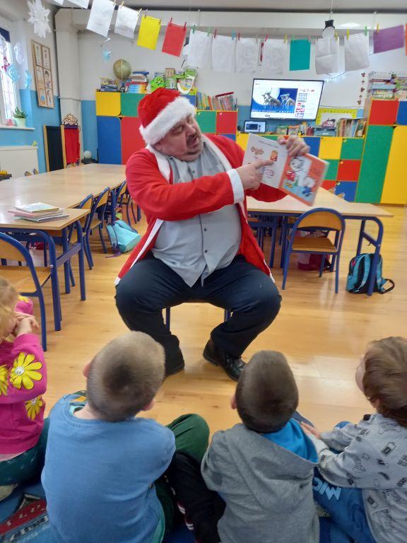 Zdjęcie przedstawia dyrektora biblioteki siedzącego na krześle w przedszkolu w stroju Mikołaja. Wokoło dyrektora siedzą na dywanie dzieci
