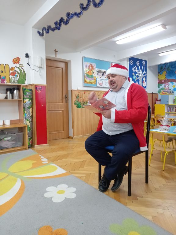Zdjęcie przedstawia dyrektora biblioteki siedzącego na krześle w przedszkolu w stroju Mikołaja. Wokoło dyrektora siedzą na dywanie dzieci. Pozując do wspólnego zdjęcia