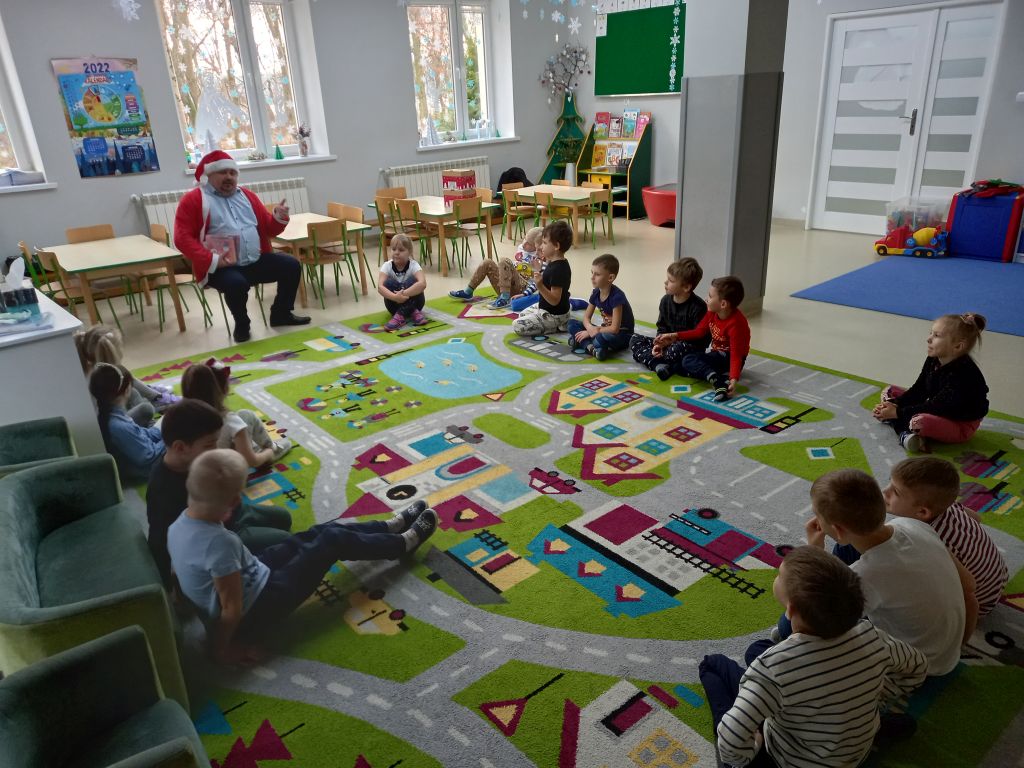 Zdjęcie przedstawia dyrektora biblioteki siedzącego na krześle w przedszkolu w stroju Mikołaja czytającego bajki