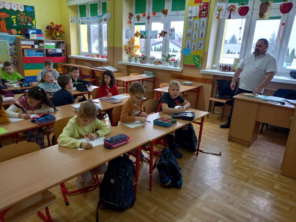 Zdjęcie przedstawia uczniów siedzących przy stolikach w szkole w trakcie lekcji. Koło dzieci stoi dyrektor biblioteki prowadzący zajęcia lekcyjne